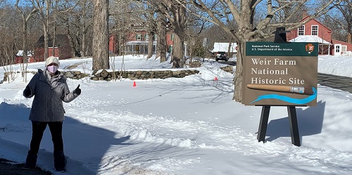 A visitor wearing a mask stands next to park sign that reads "Weir Farm National Historic Site."