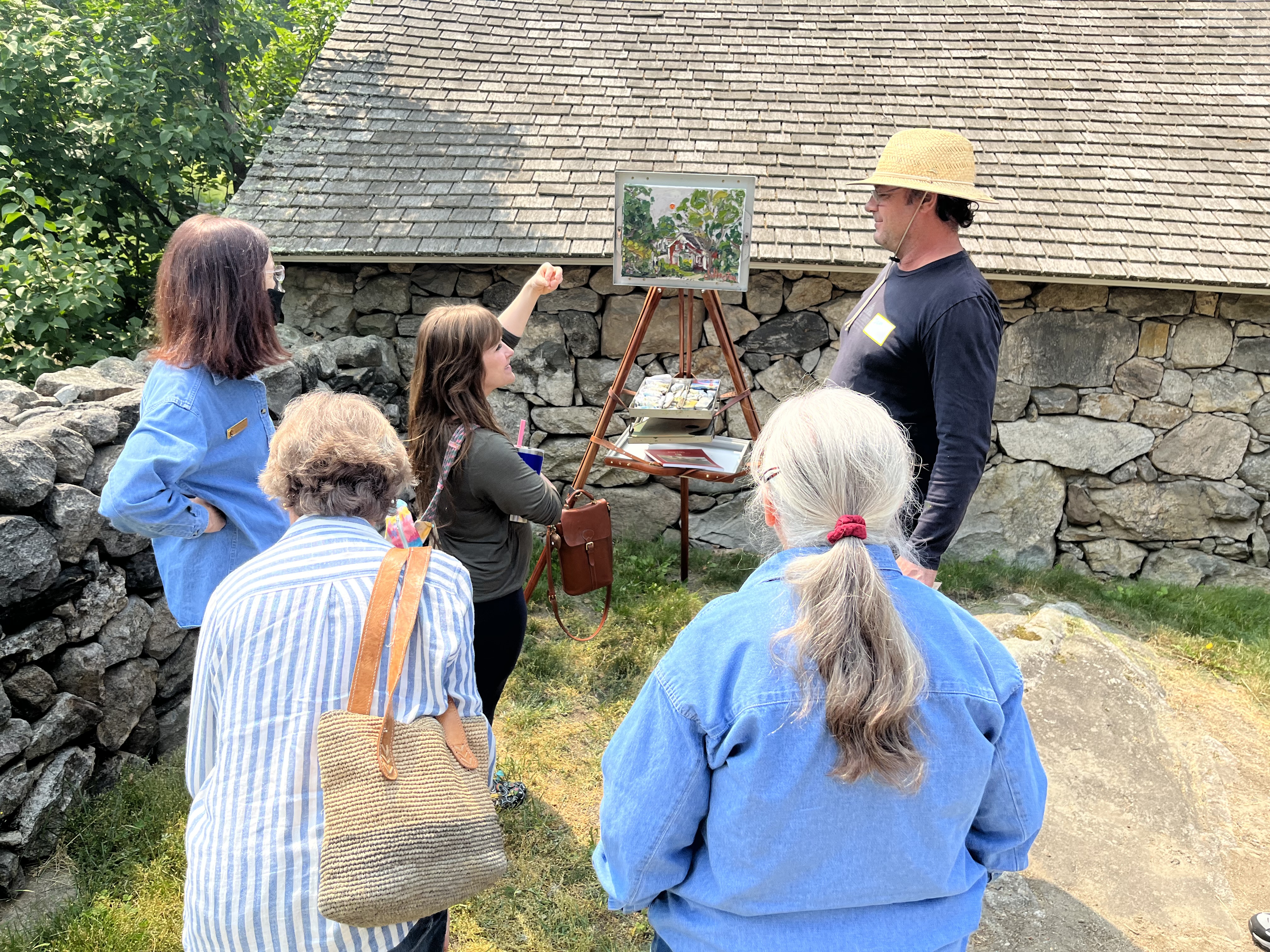 A group of people outside stand around a painting easel talking