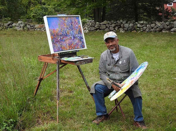 A man sitting in front of an easel in a meadow, holding a paint brush and a pallet.