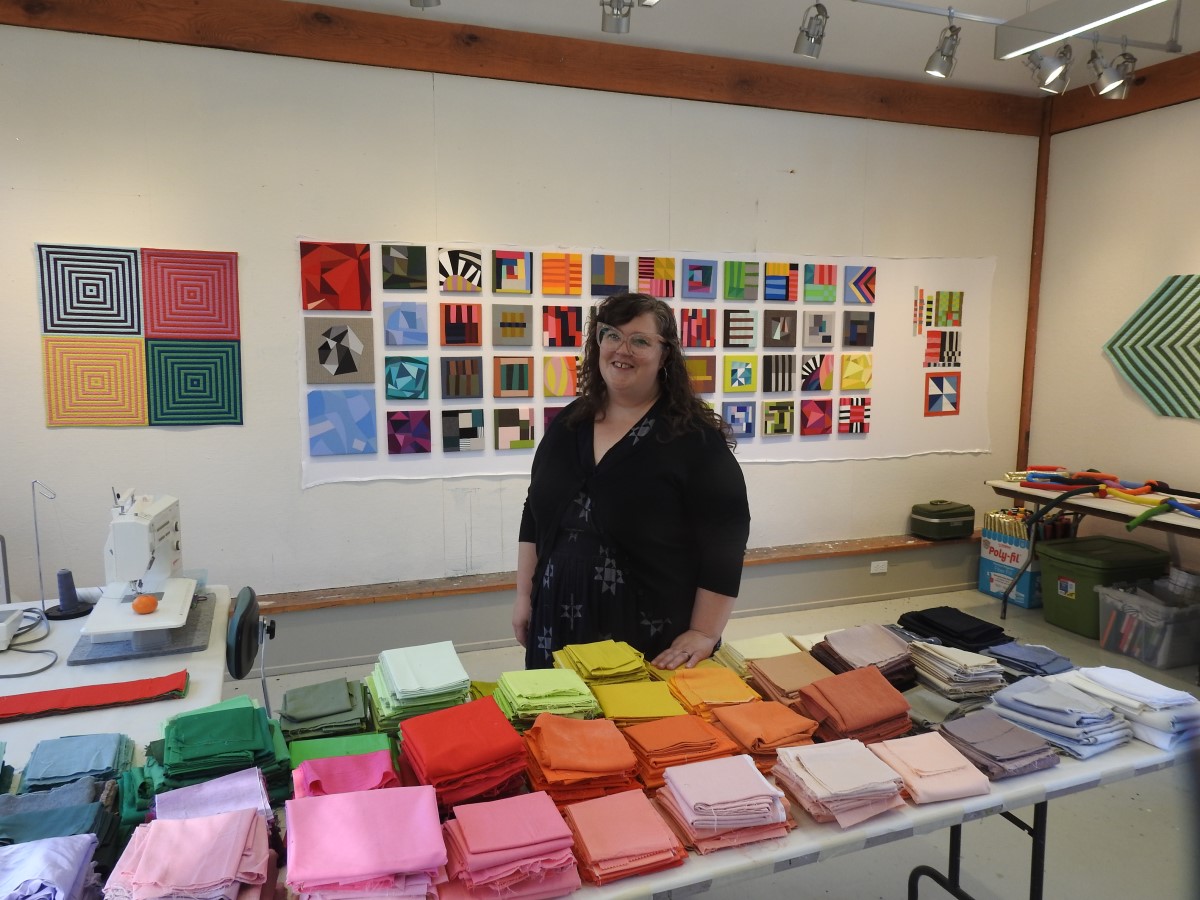 A person stand behind a table covered in colorful fabric squares neatly piled by color. Behind her on the wall are dozens of small, square works of art in colorful, geometric patterns.