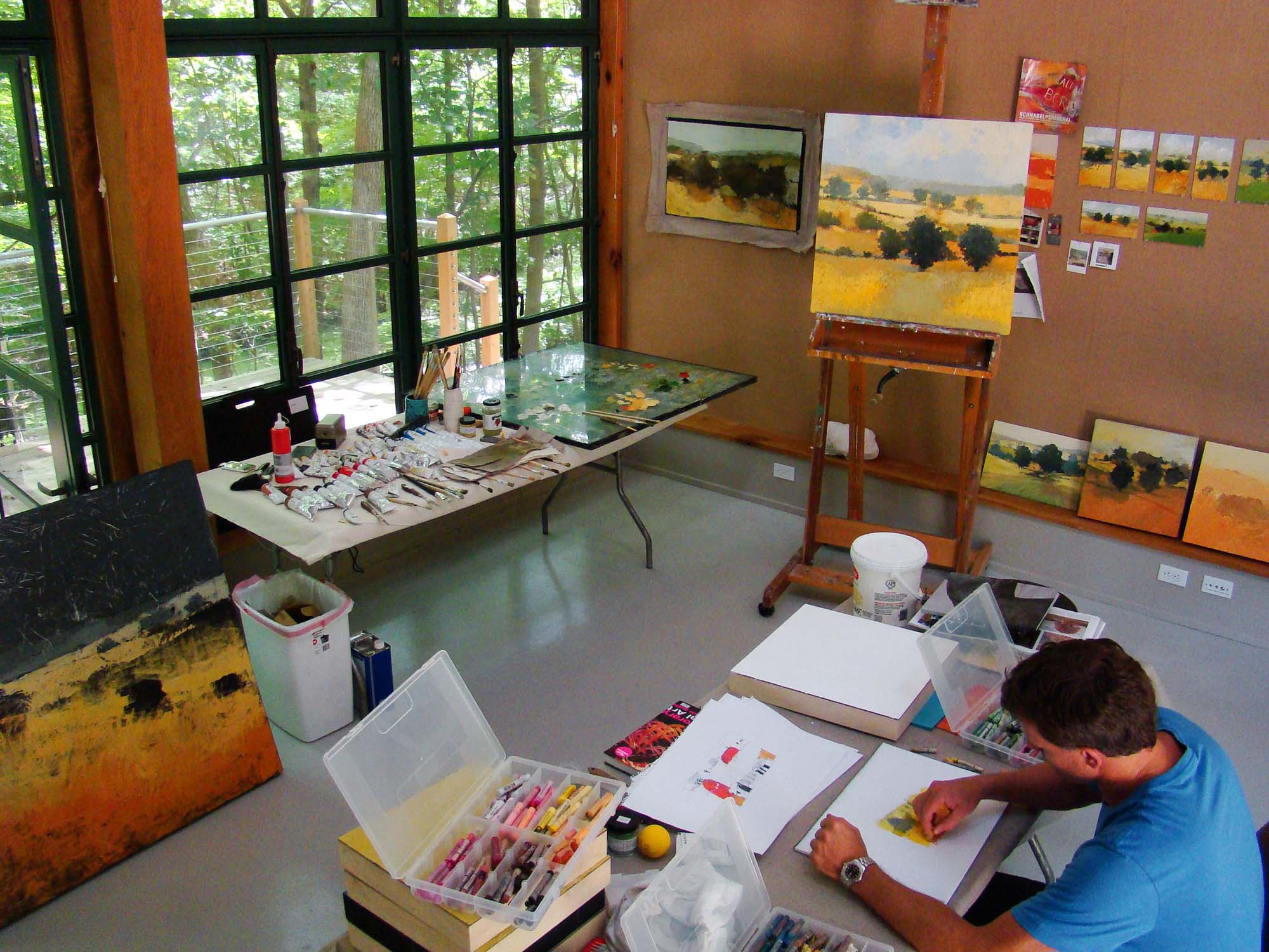 A modern art studio filled with paintings of landscapes. An artist sits on one side working on a drawing with pastels.