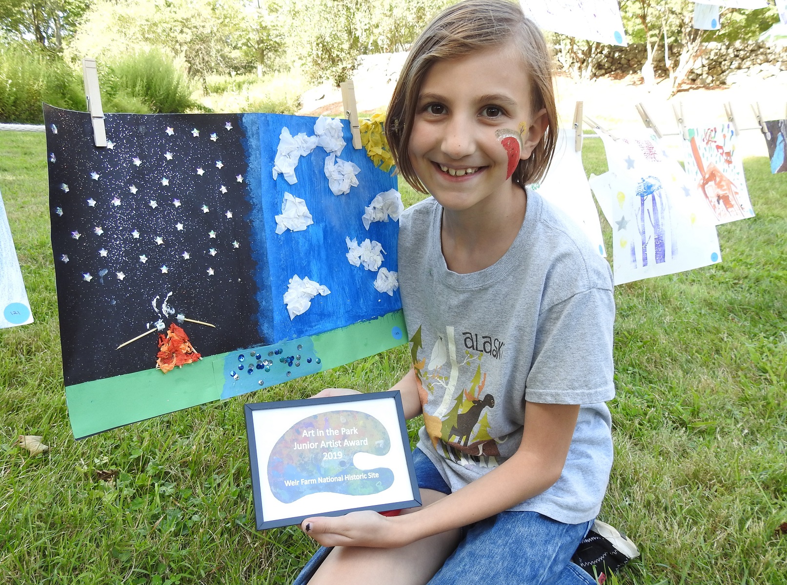 A girl kneels next to a clothesline filled with art. In her hand is a small black picture frame.