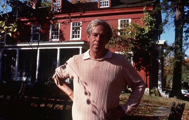 A man with grey hair, wearing a light brown sweater, standing in front of a red house with white trim.