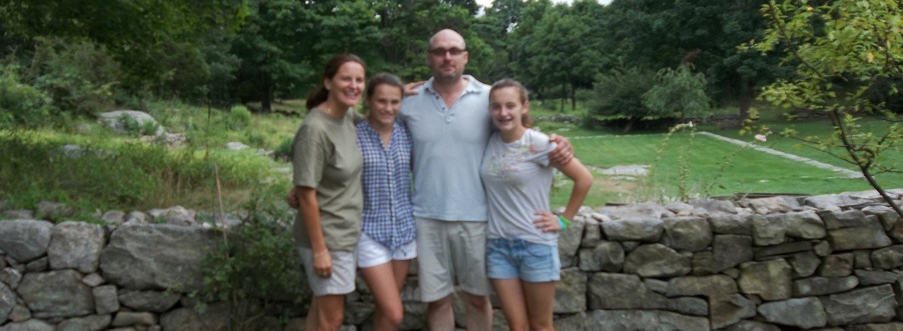 A family of 4 standing in front of stone wall with trees and garden in background at Weir Farm during a permitted photo session