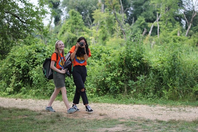 Two girls holding cameras walk on a trail.