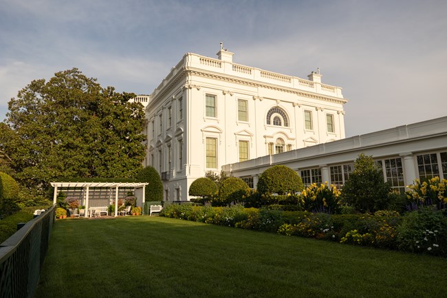A rectangular mowed lawn with a pergola at the rear and a garden of flowers by the White House.