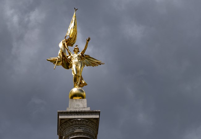 A statue of a golden, winged person holding her hands aloft with a flag in one, and the other hand open.