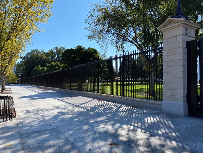 Tall black iron fence separates a paved walkway from the White House lawn.