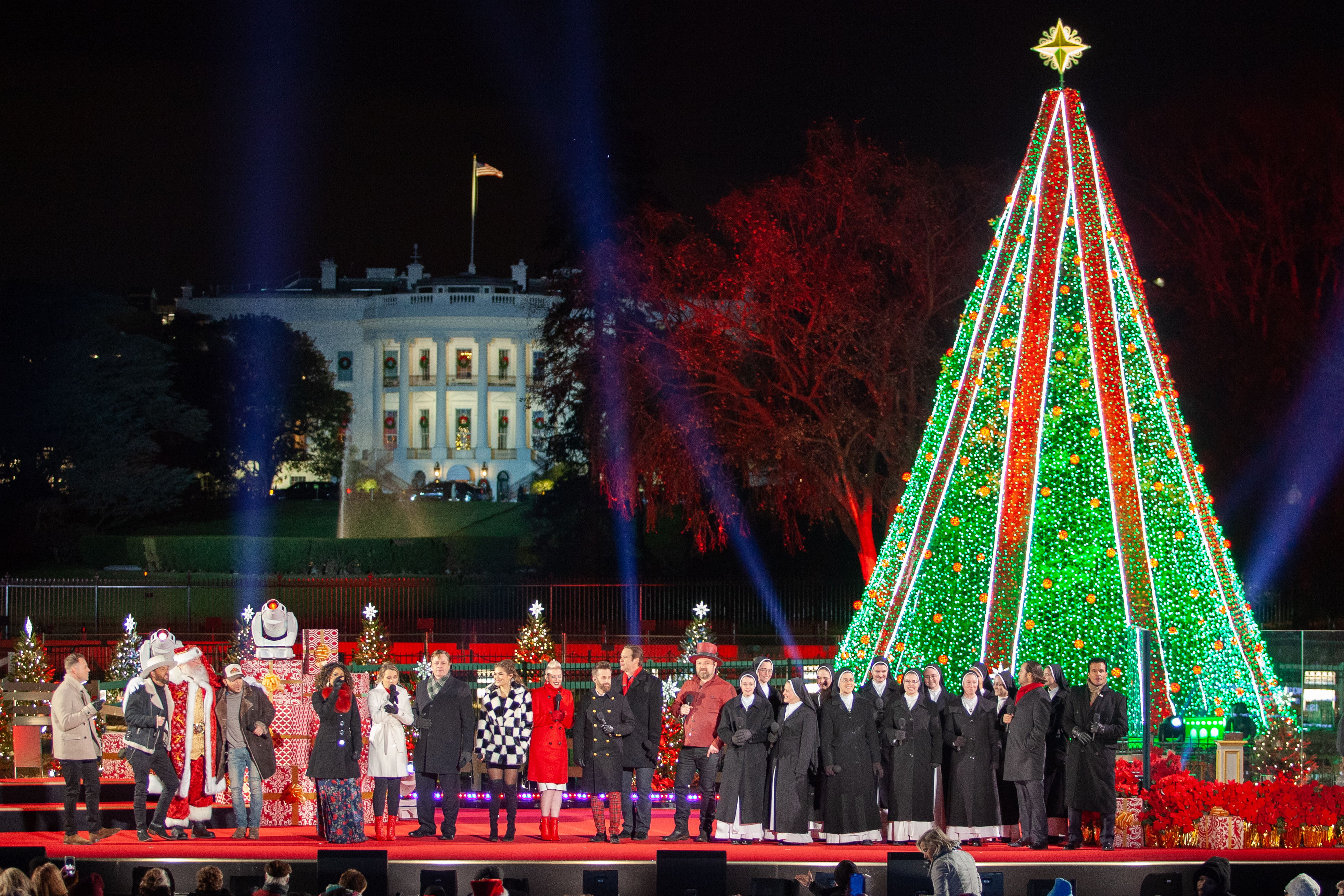 Talent sing in front of the lit 2018 National Christmas Tree with the White House in the background.