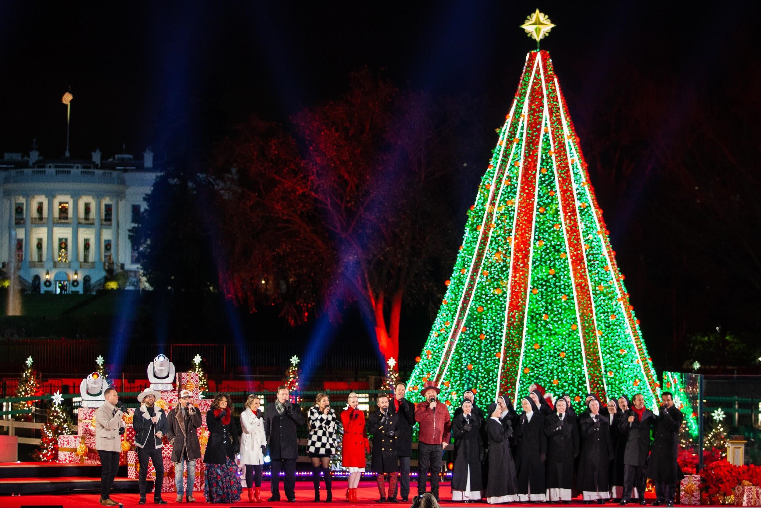 Singers sing in front of a towering, lit Christmas Tree.