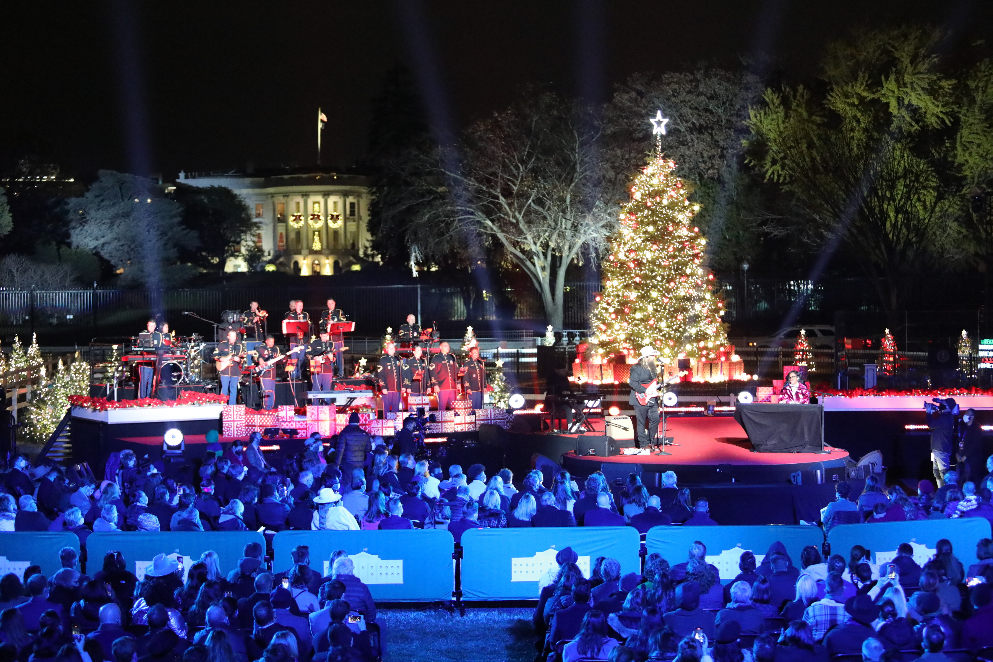 The National Christmas Tree is lit with the White House in the background. The forefront of the image is the stage with performers and the audience.
