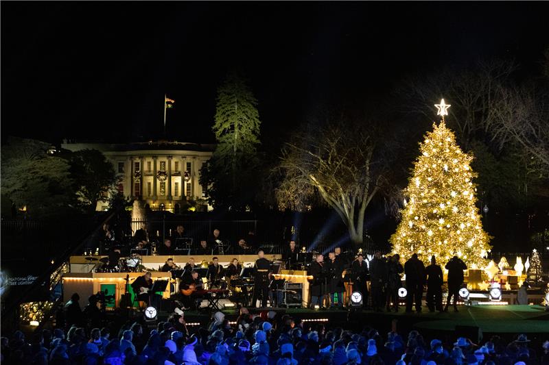 2022 National Christmas Tree Lighting - 2024 News Release Stage with a tall lit up Christmas tree with a band playing with the South view of the White House behind. An audience cheers on under blue lights.