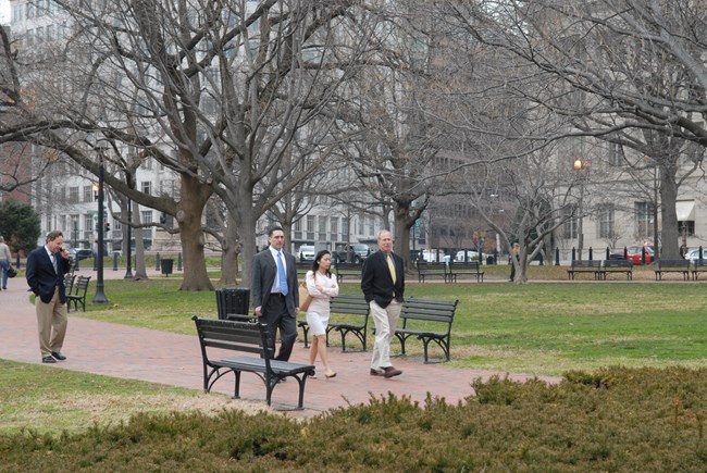 Three people in a park walk on a wide brick sidewalk lined with benches.