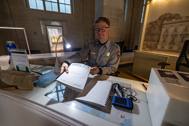 A ranger sitting at a desk holds a booklet with braille.