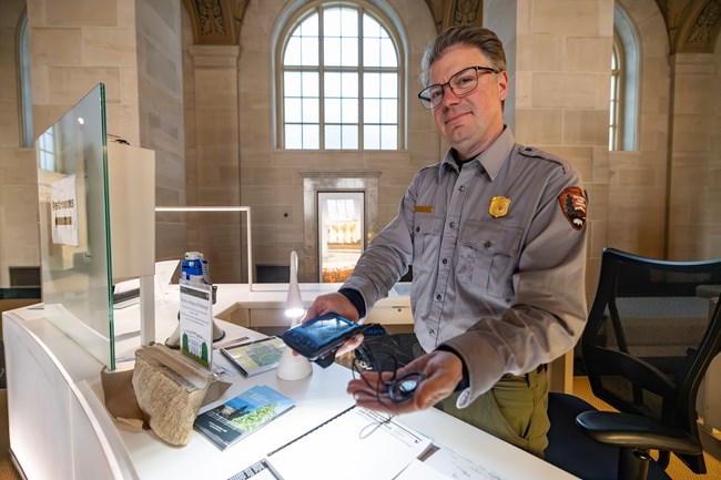 A ranger standing behind a desk holds an assistive listening device with earpiece.