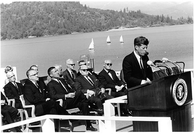 Black and white photo of President John F. Kennedy standing at podium dedicating Whiskeytown Dam in September 1963. Whiskeytown Lake and sailboats in background. Large crowd in front of Kennedy.
