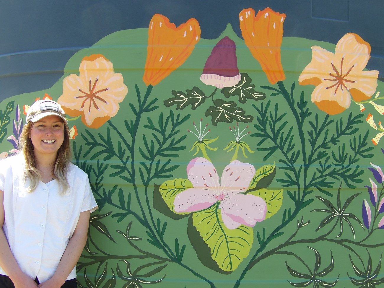 White female with long blonde hair and ballcap and white shirt standing in front of painting depicting orange and pink flowers and green leaves.