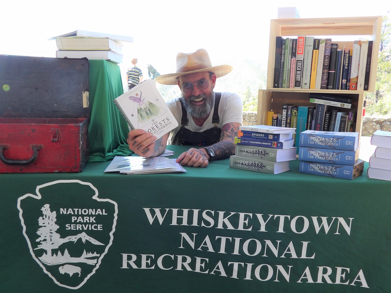 Obi Kaufmann, with beard, straw hat, suspenders, and white shirt, smiling at camera and holding up his book. Obi is sitting behind a table with a green table cloth and several books are on top of the table.
