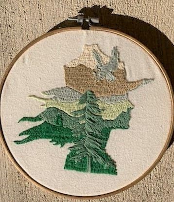 Artistic weaving showing a female park ranger with brown and beige flat hat, face, green hair and shoulders. Tree, bird, and mountain weaved into it.