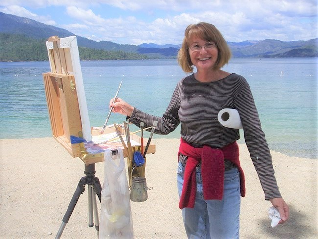 A female artist painting and smiling. Whiskeytown Lake in background.
