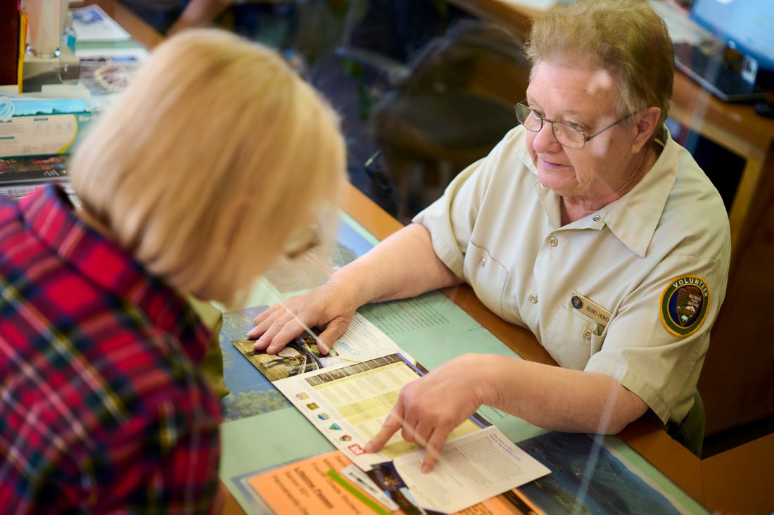 A white female senior citizen in volunteer shirt assisting a visitor at the Visitor Center.