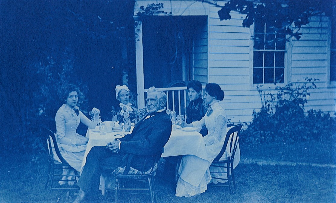 Black and white photo of Charles, Philena, and three daughters, sitting around a table in front of the Camden House.