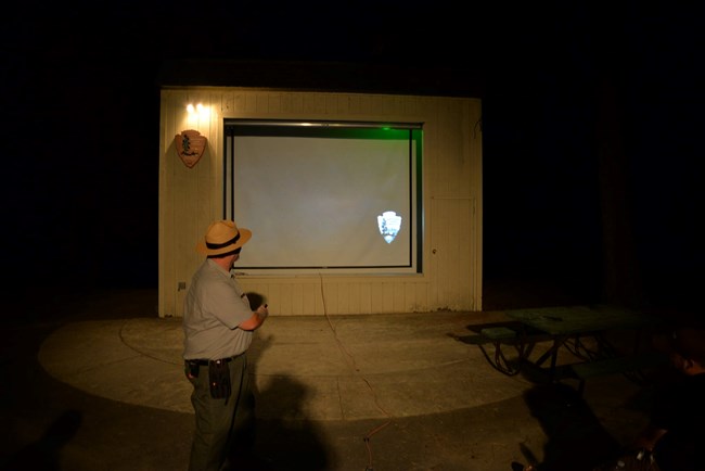 An interpretive park ranger in green and gray uniform and flat hat in front of power-point slide at Oak Bottom Amphitheater. Dark black background spotlighting the night.