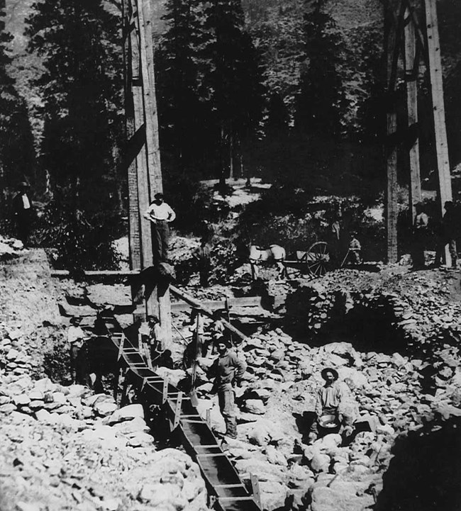 Black and white photo of goldminers and goldmining operations along Whiskey Creek. Long Tom, panning, field of rocks.
