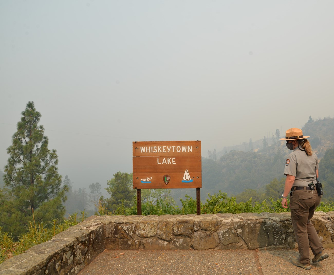 A female park ranger in flat hat, green pants, and grey shirt looks out over very smoky skies and low visibility. She should be able to see Whiskeytown Lake from her vantage point, but she can't because of the smoke.