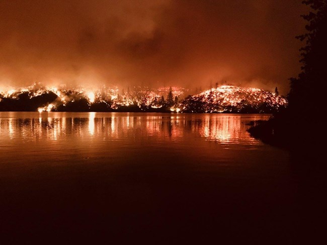 Carr Fire and Whiskeytown Lake at night. Large red and orange flames rising in the woods beyond the lake.