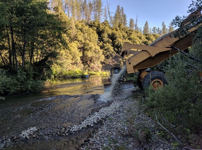 A large yellow dump truck dumping gravel into lower Clear Creek.