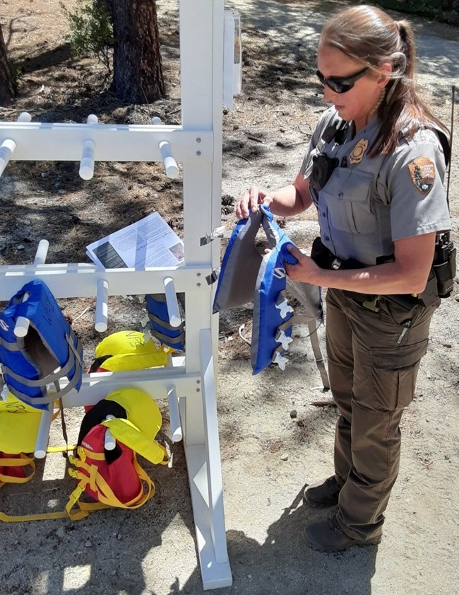 A Whiskeytown law enforcement park ranger holding a youth lifejacket and standing next to a rack of youth lifejackets at Brandy Creek Beach. The lifejackets are red and blue.