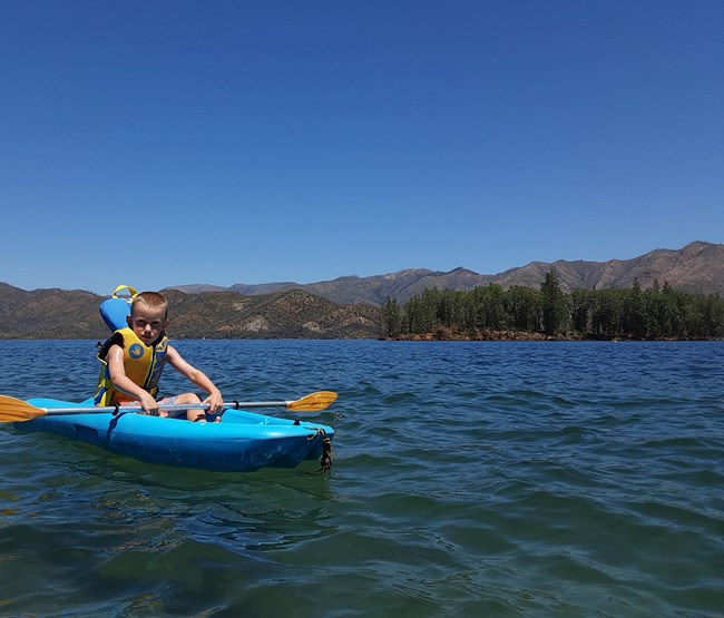 A Caucasian boy on a youth kayak holding a paddle on Whiskeytown Lake. Blue water and blue sky.