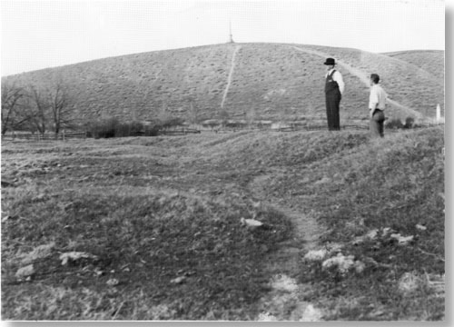 Historic photo of two men standing on small rise looking down. There is no water. Vegetation is very sparse.