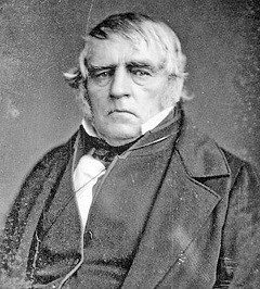 A black and white photo of a man from the stomach up. He is dressed in a suit and vest and a high white collar. His hair covers his ears and is white.