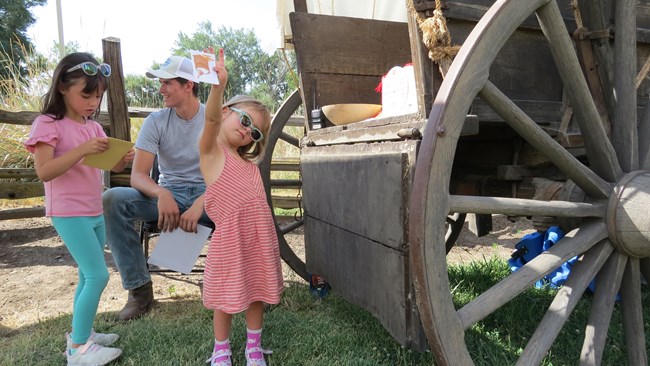 In the middle of the frame, a young girl holds a piece of paper with a flour sack on it to the camera while another holds a bag and an employee sits behind them, the rear of a wagon and wheel covers the right side of the frame.