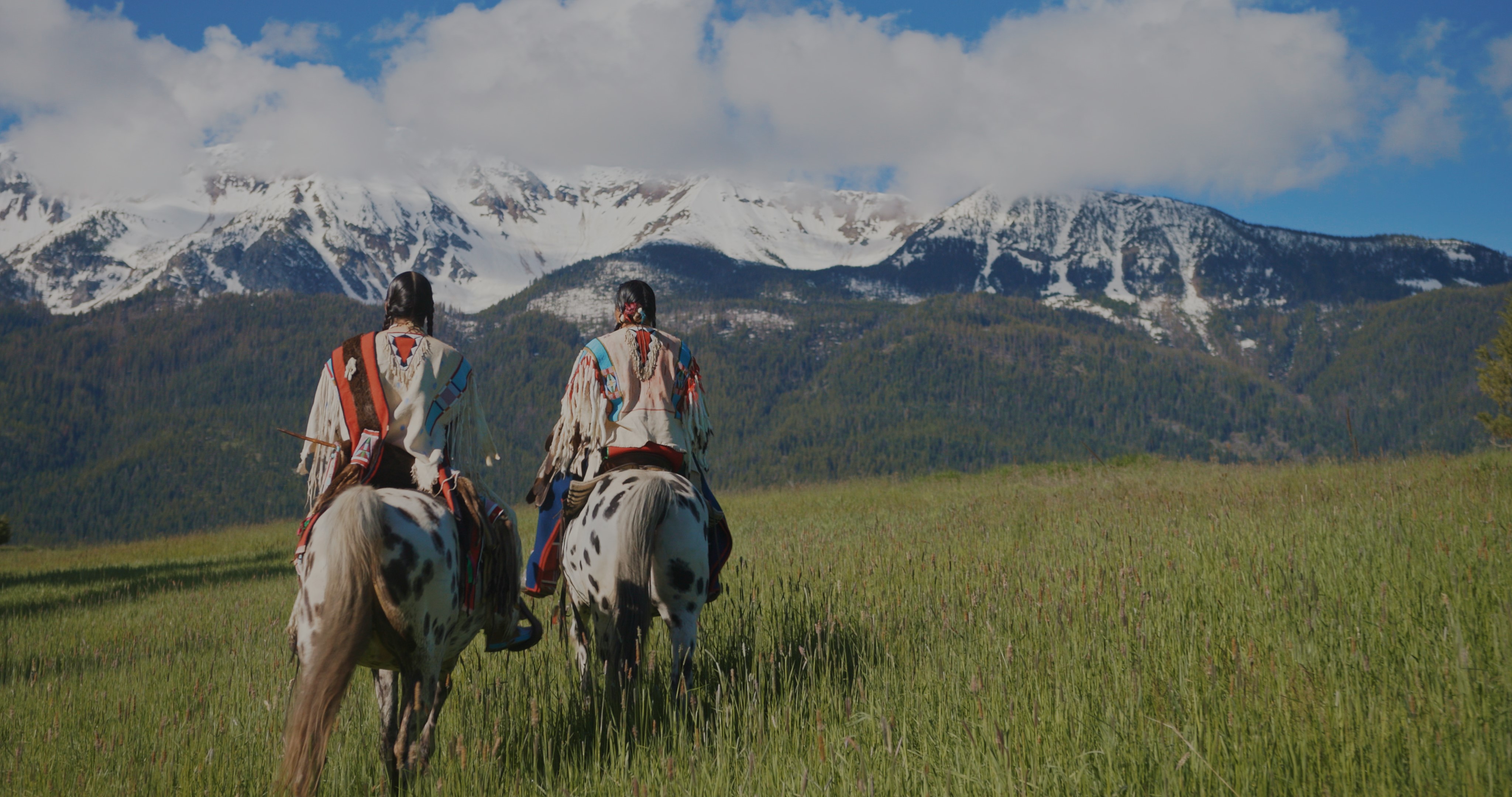Covenant of the Salmon People Two people on white spotted horses look at a snow covered mountain. The horses stand on a field of green grasses