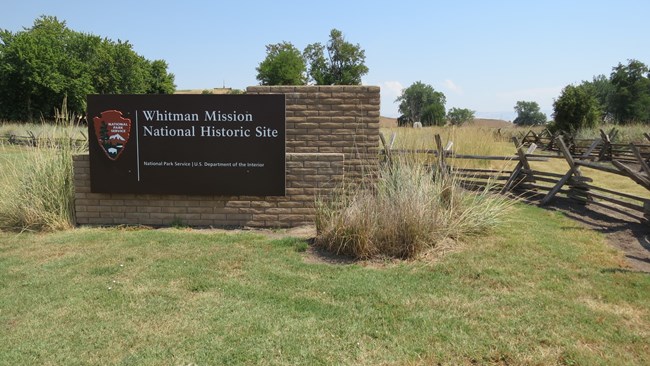 A brown sign with the text "Whitman Mission National Historic Site" and the NPS arrowhead to the left of the text, behind the sign is tall grass fields and split-rail fences
