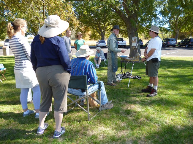 A group of people outside listening to a man talk while painting