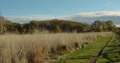 A field of tallgrass next to a split rail fence, all under blue skies. A hill with an obelisk monument on top is centered in the background