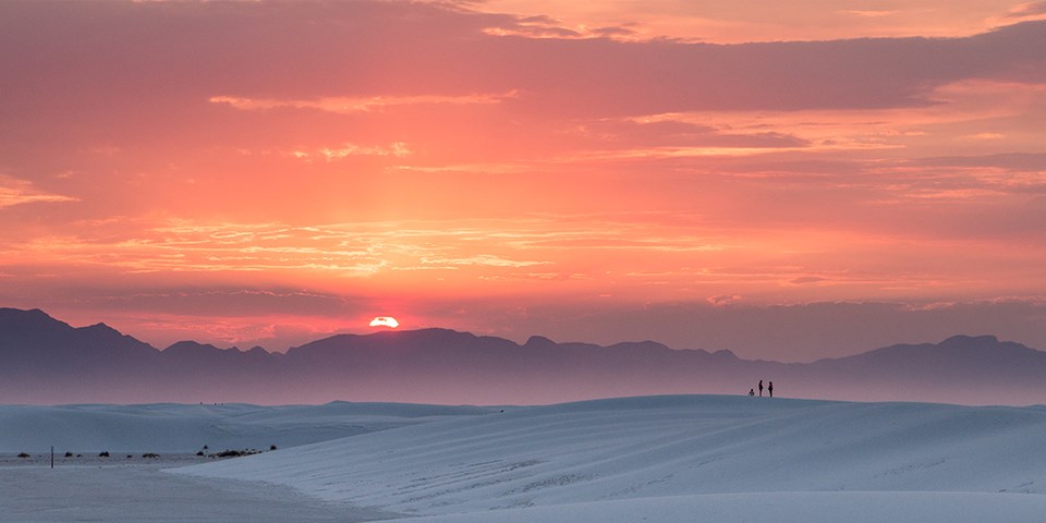The sun sets amidst a pink and orange sky above the dunes