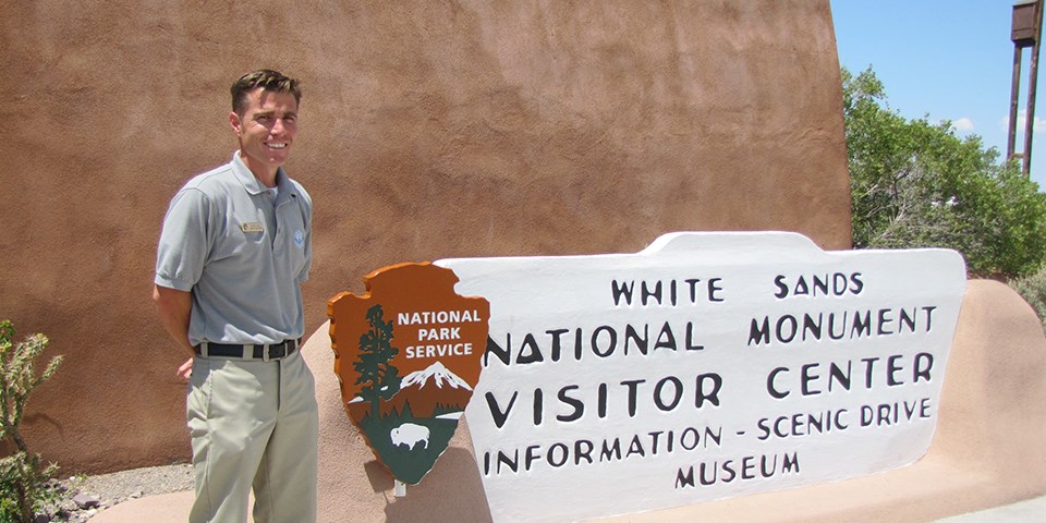 A teacher stands next to a park service arrowhead and visitor center sign.