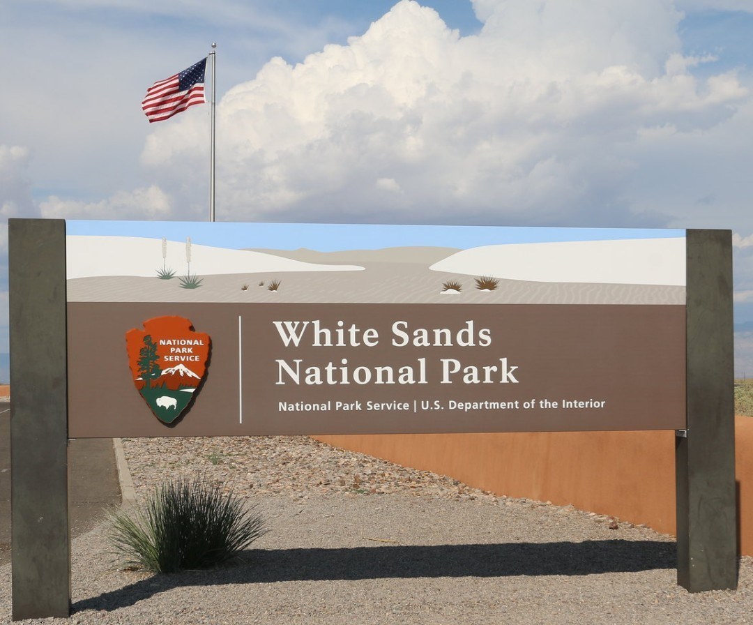 a sign saying White Sands National Park with a artistic rendition of White Sand dunes and a roadrunner. An American flag flies on a flagpole behind the sign.