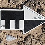 A black and white arrow in brown dirt to provide scale for the adjacent stone arrowhead