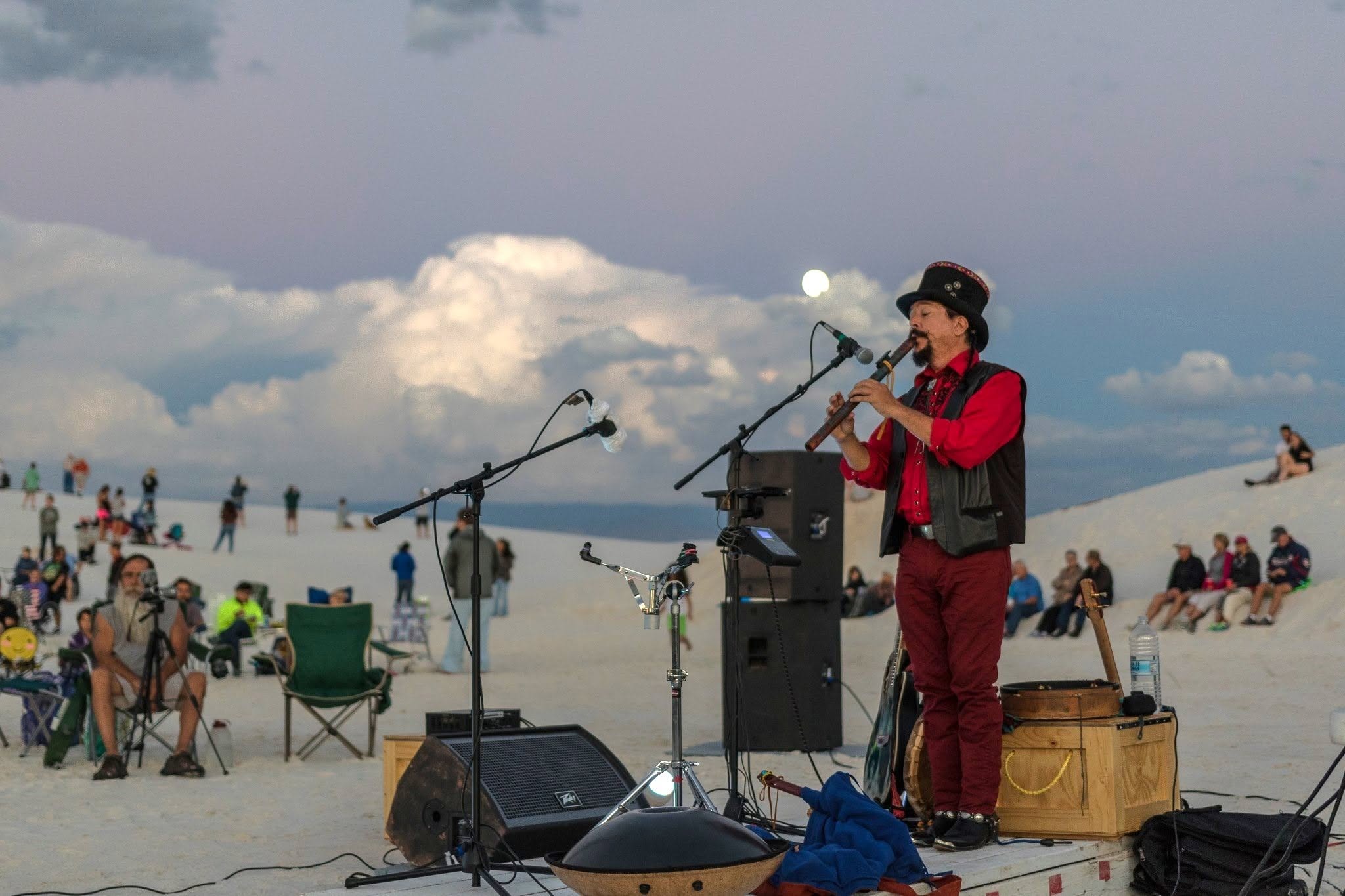 A musician in a red shirt and top hat plays a flute on a stage set upon white sand dunes, with an audience watching from the background under a cloudy, evening sky.
