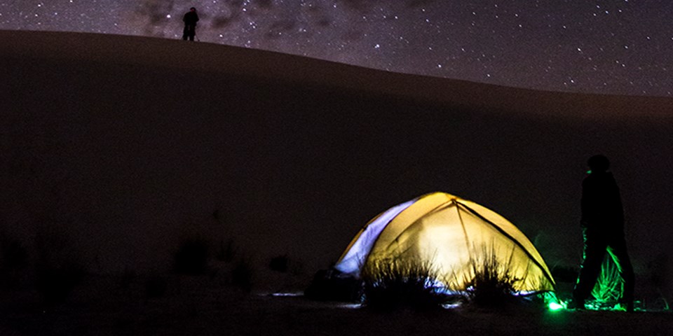 Two visitors camping on the dunefield, one is on the left far away on top of a dune and the other is on the foreground next to a tent.