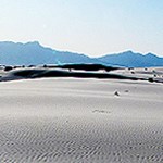 White sand dunes with ripples, mountains in the background, and blue sky