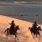 Two riders on horses galloping through sand