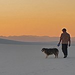 Man walking a dog on white sand with an orange sunset