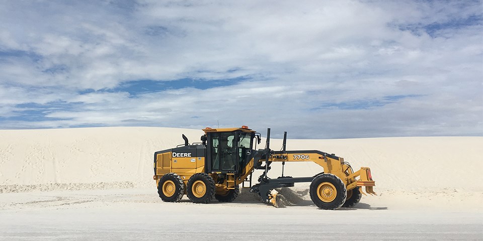 A road grating machine rides along the burm of a sand dune